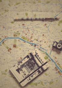 Various travel-related items are laid out on a map. The objects include a small model of the Eiffel Tower, a vintage camera, a modern instant camera, a notebook with a sketch of an architectural landmark, and a pencil. The map shows detailed streets and locations, likely from a city in Europe.