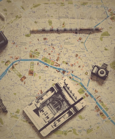 Various travel-related items are laid out on a map. The objects include a small model of the Eiffel Tower, a vintage camera, a modern instant camera, a notebook with a sketch of an architectural landmark, and a pencil. The map shows detailed streets and locations, likely from a city in Europe.