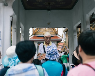 A group of tourists are gathered around a guide who is holding a brochure or map. The setting appears to be a historical or cultural site with ornate architecture in the background and an archway. The guide is wearing a hat and glasses, and the tourists are mostly wearing casual clothing and hats.
